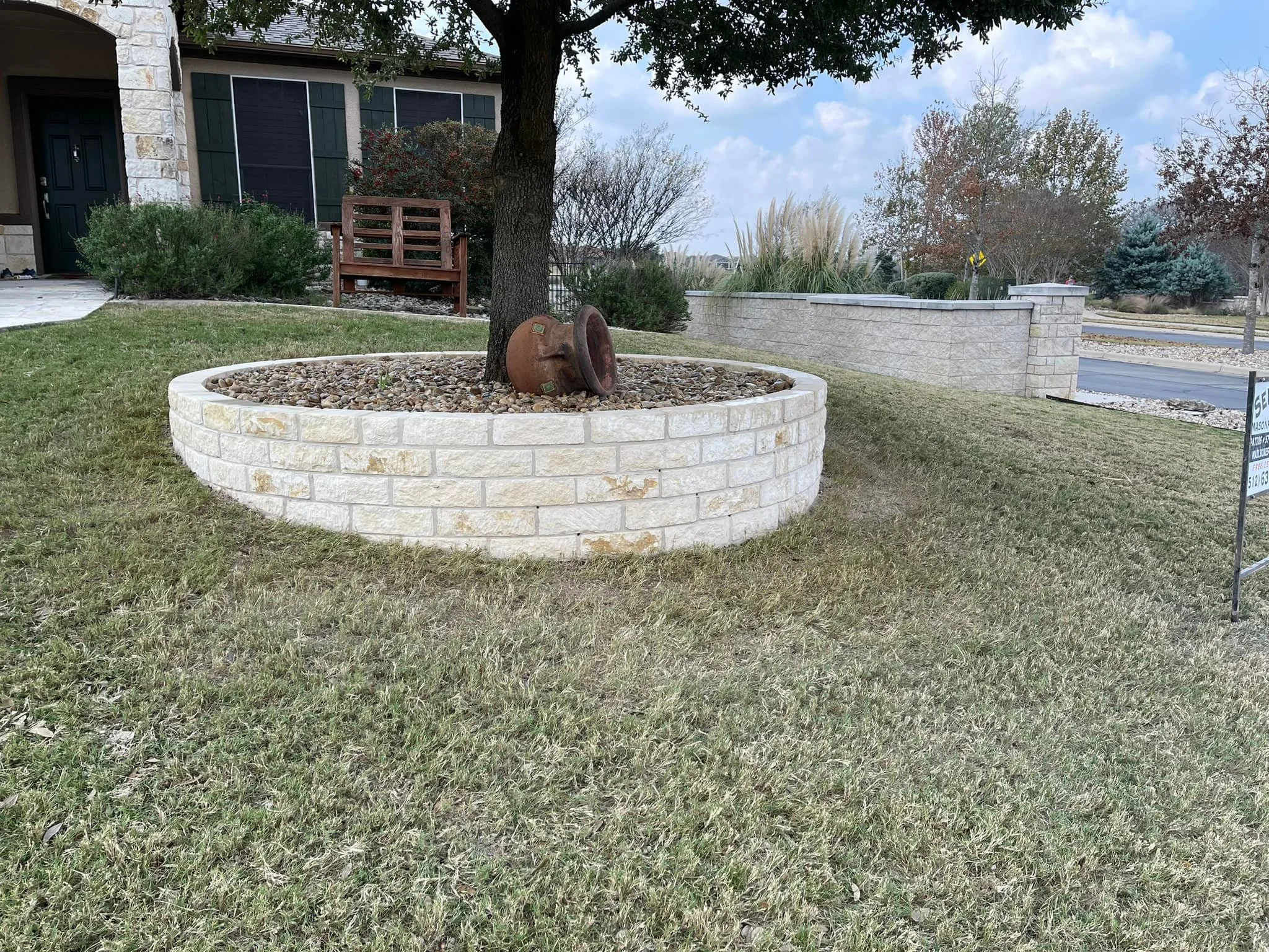 A front yard with a circular white brick planter containing a tree and a rusty, overturned clay pot. There is a house with a stone and siding exterior, a wooden bench, and some bushes in the background. The lawn is green with some brown patches, and 