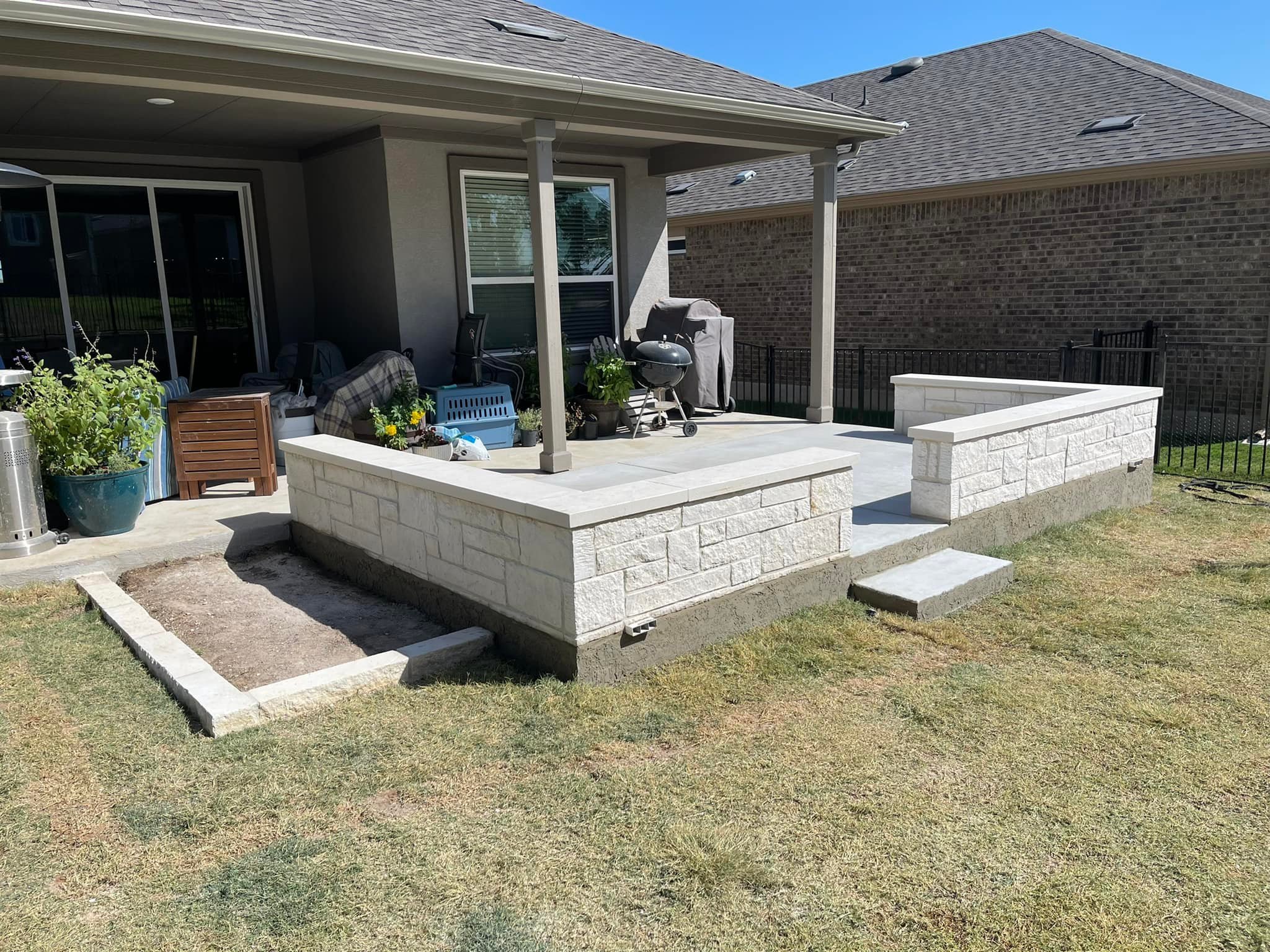 Backyard patio under construction with white stone wall, concrete flooring, steps, and a covered porch area with outdoor furniture, grill, potted plants, and gardening tools.