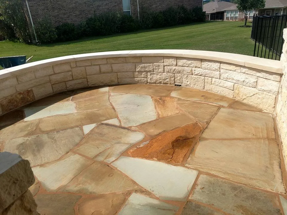 Backyard patio with cobblestone and stone pavers, surrounded by a black metal fence, with green lawn, bushes, and houses in the background.