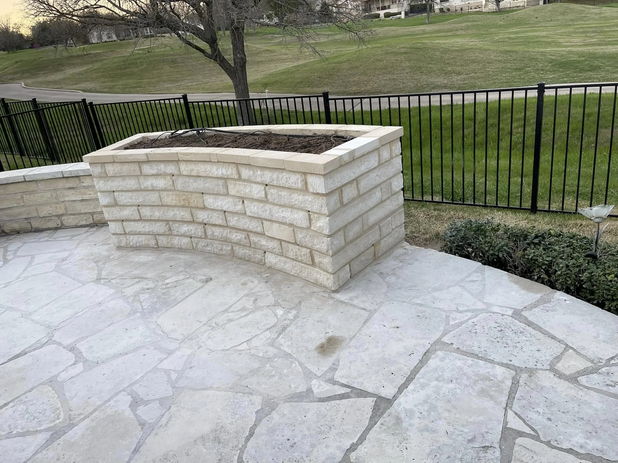 A curved stone planter with soil on top, located on a light-colored stone patio, in a backyard with a black metal fence, grass, trees, and a golf course hill in the background.