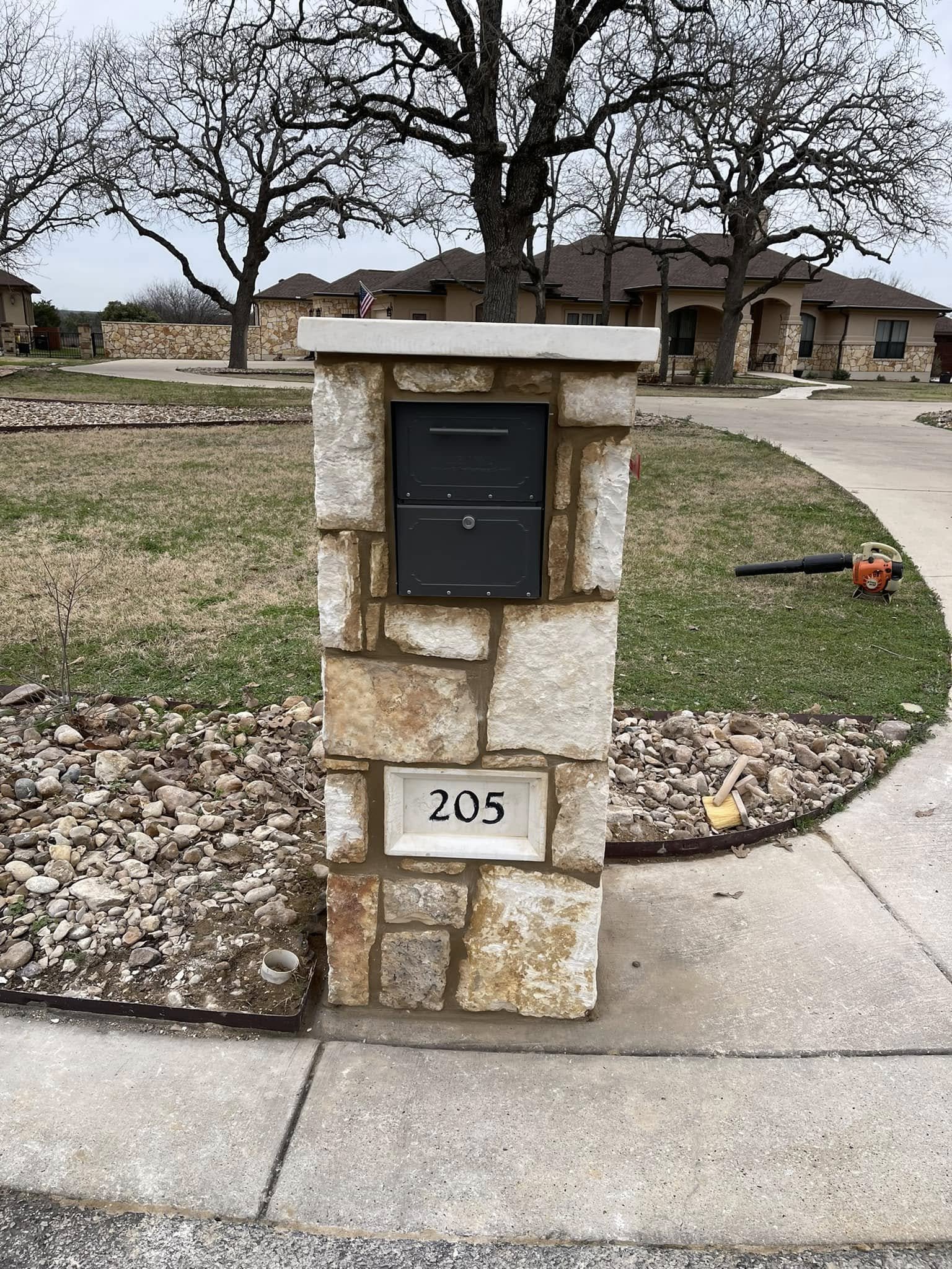 Stone mailbox with house number 205 and a black mailbox mounted on a stone pillar in front of a suburban house with trees and a driveway.