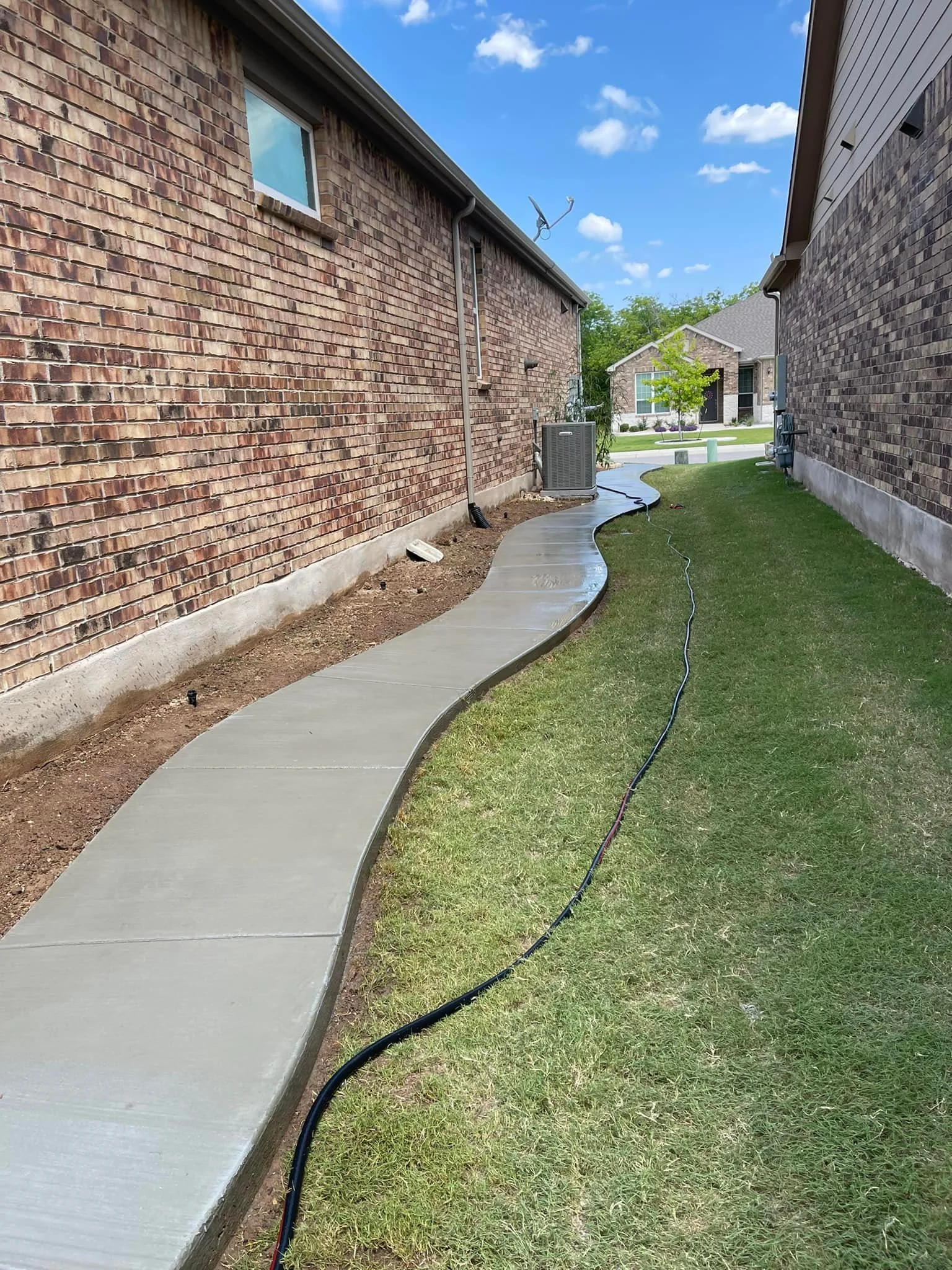 Newly poured concrete sidewalk with a wavy shape runs between two brick houses under a blue sky with clouds. A garden hose is lying on the grass beside the sidewalk, and an air conditioning unit is visible on the front of one house. A small tree and 