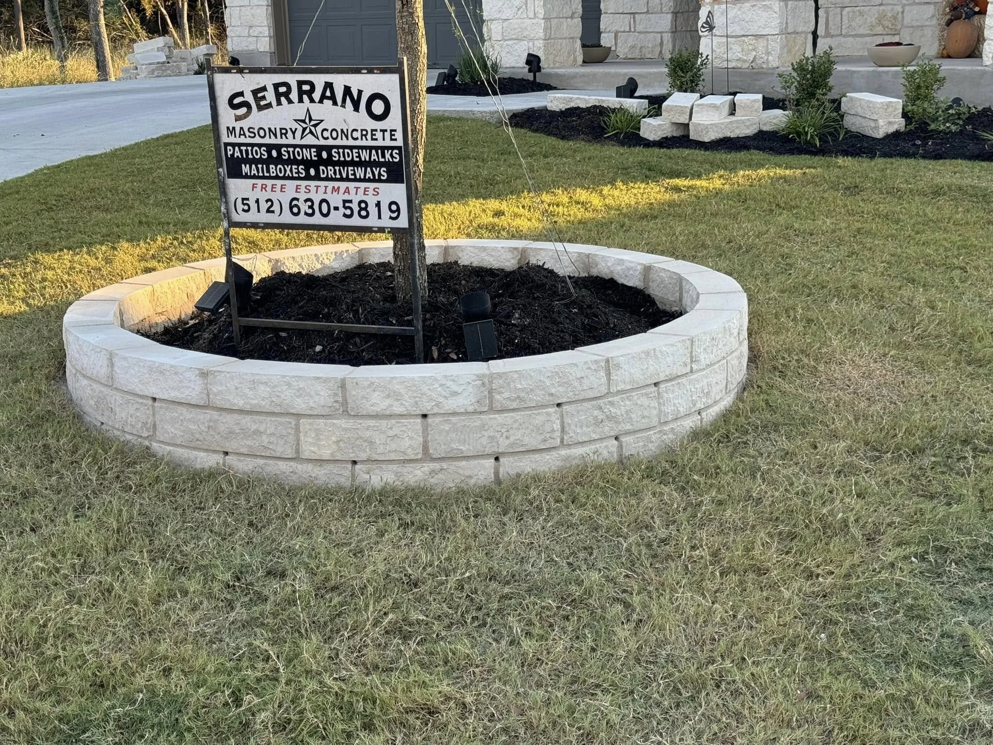 A circular flower bed with white brick edging, filled with soil and a sign for Serrano Masonry & Concrete, located in front of a house with a small lawn and plants.