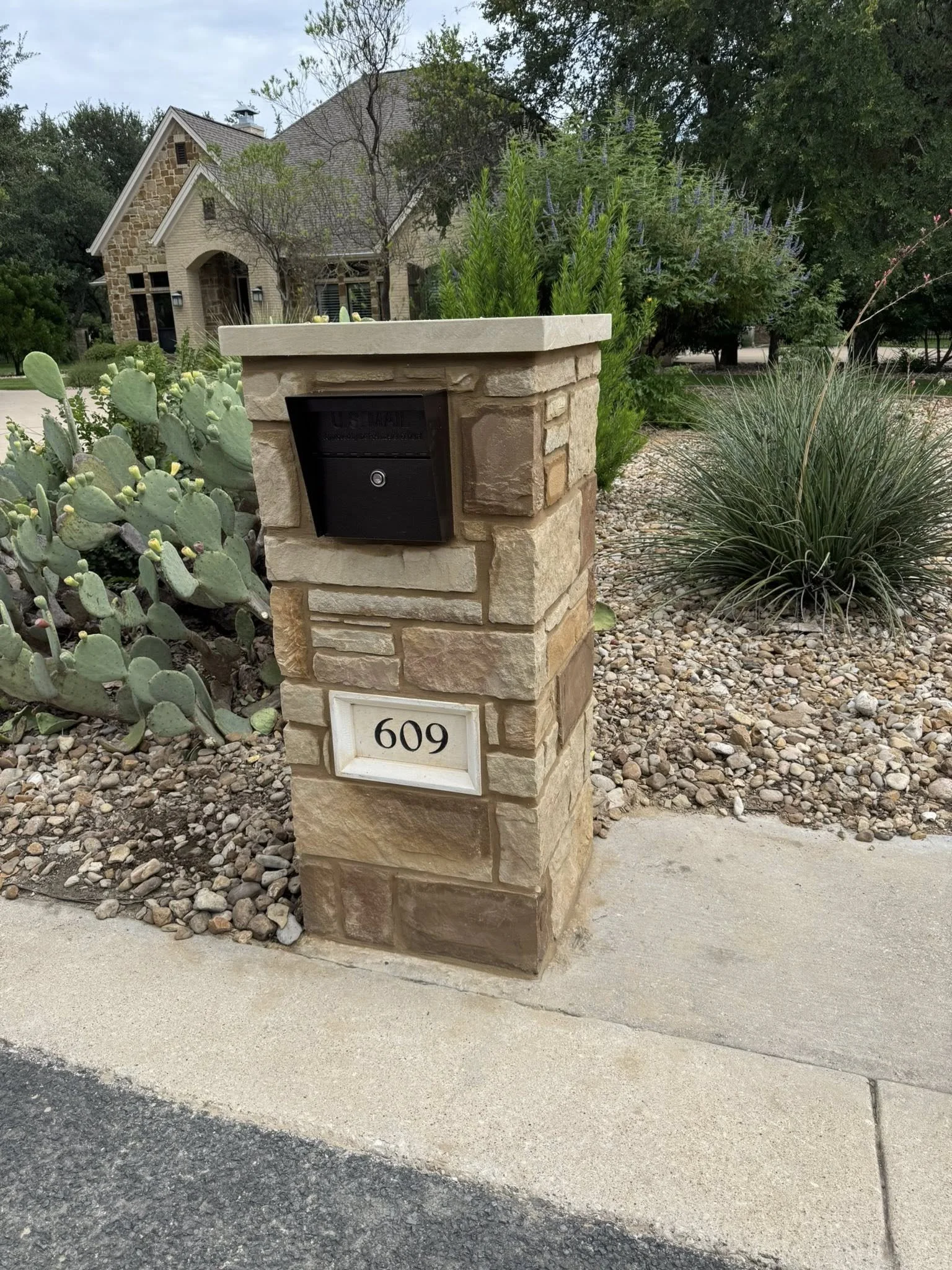 A stone mailbox with the house number 609 on it, situated at the edge of a sidewalk with decorative rocks and desert plants surrounding it. A house with a stone exterior and gabled roof is visible in the background.