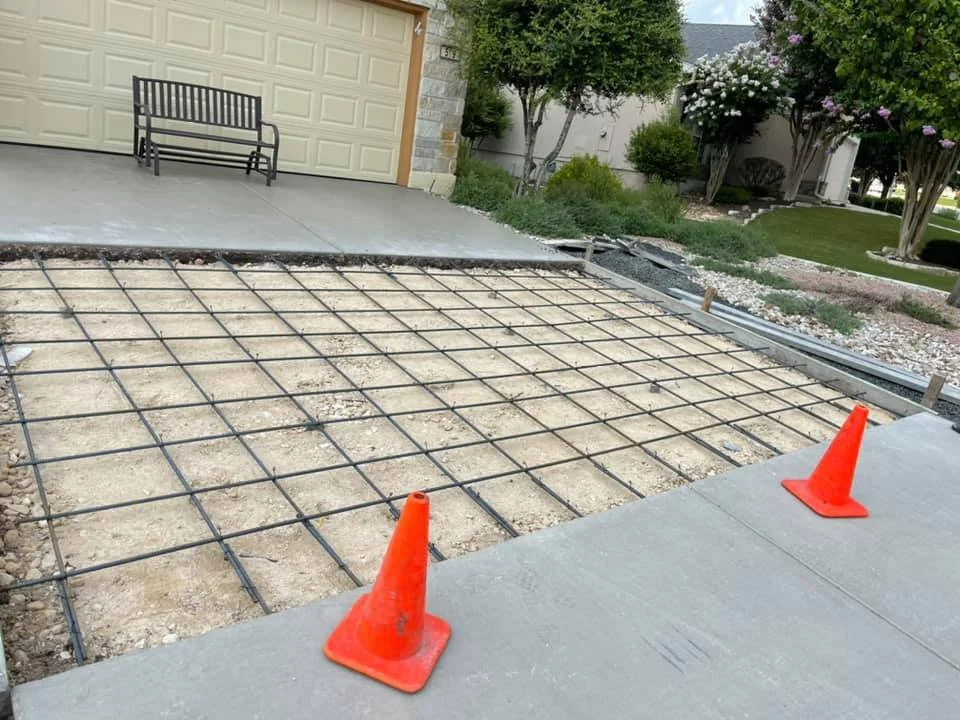 Construction site with rebar grid laid out for new concrete driveway, orange traffic cones marking the area, and a finished concrete driveway in the foreground.