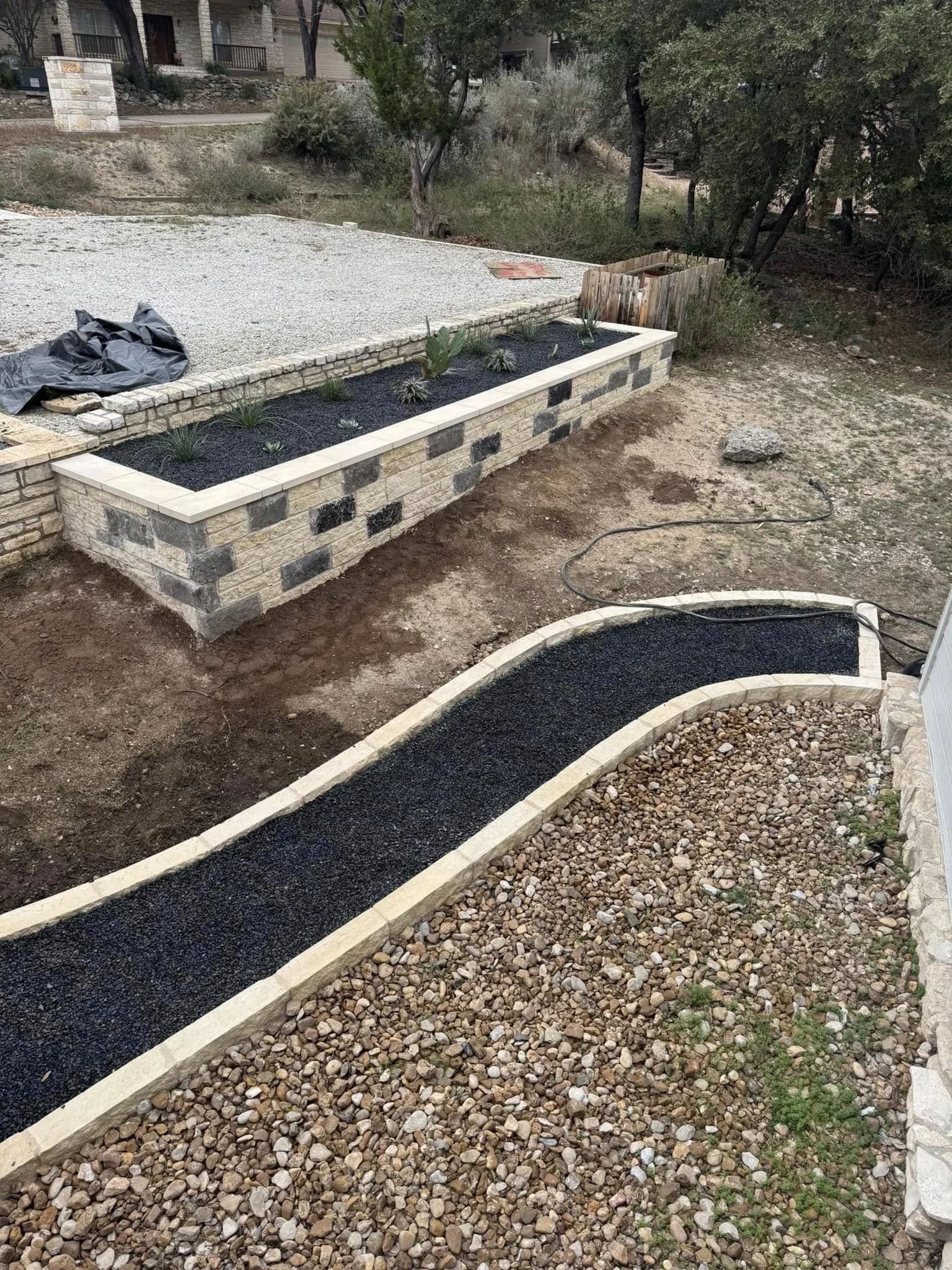 A landscaped backyard featuring a brick planter with plants, a curved gravel pathway, and a gravel and dirt yard area, with trees and houses in the background.