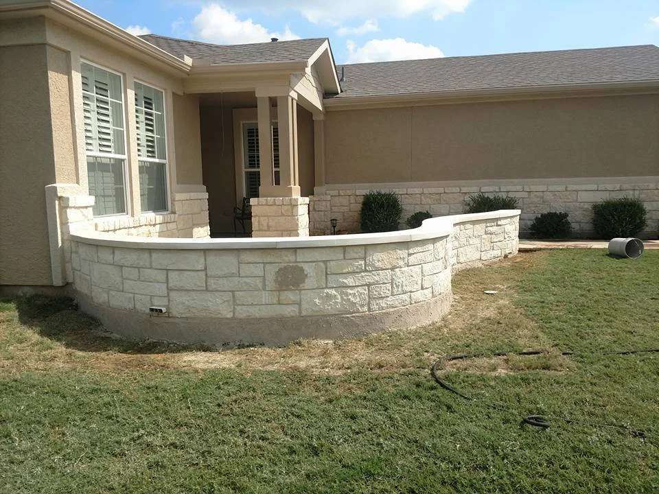 Partial view of a house with a newly built curved stone wall and green lawn. There are windows with shutters, a porch with columns, and bushes along the house.