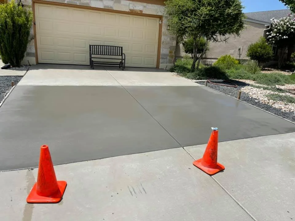 Newly poured concrete driveway with orange safety cones, a black bench, and a tree in front of a house with a light-colored garage door.