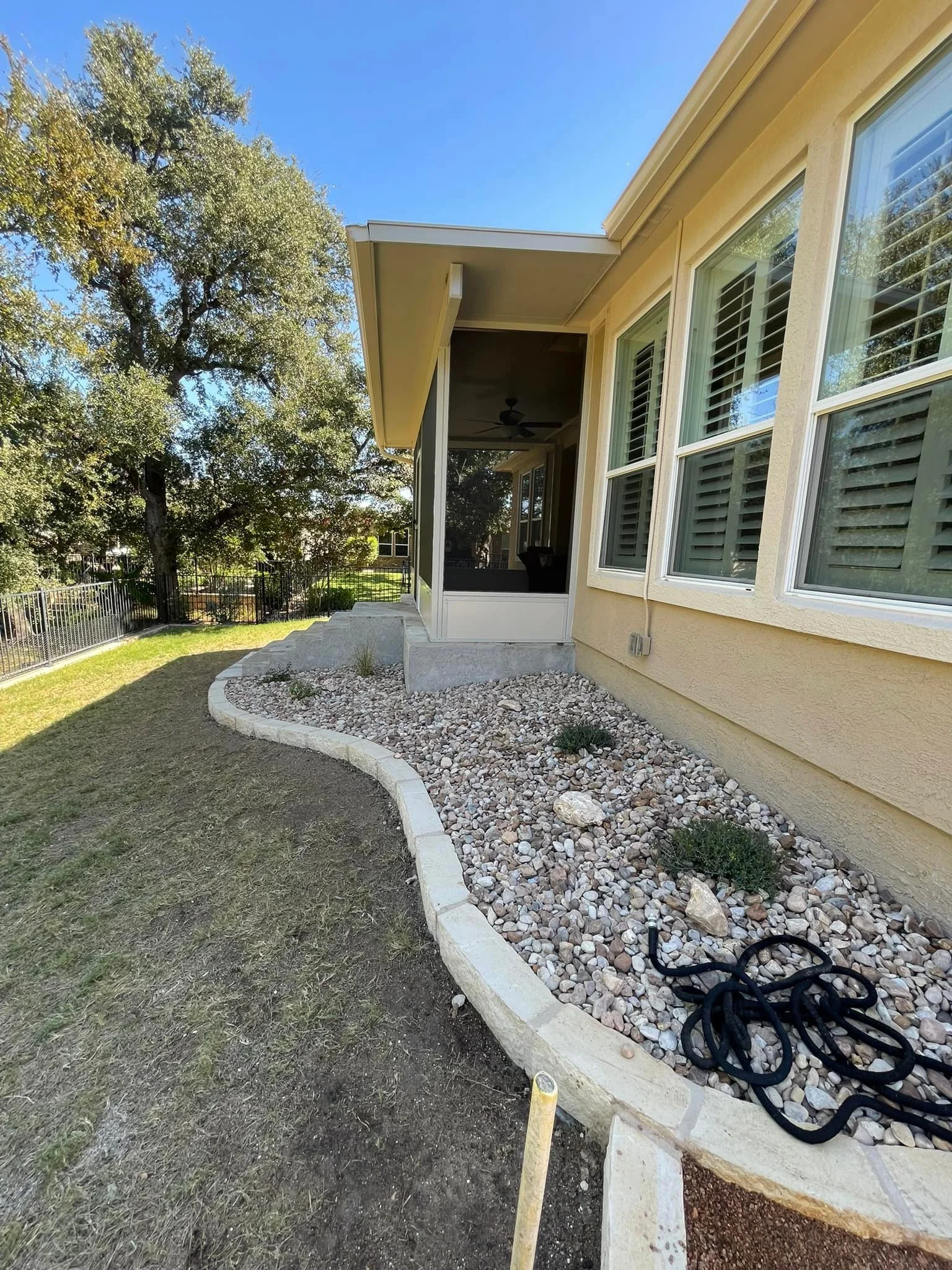 Backyard with a curved rock garden bed, beige house with large windows, screened porch, on a sunny day with a large tree in the background.
