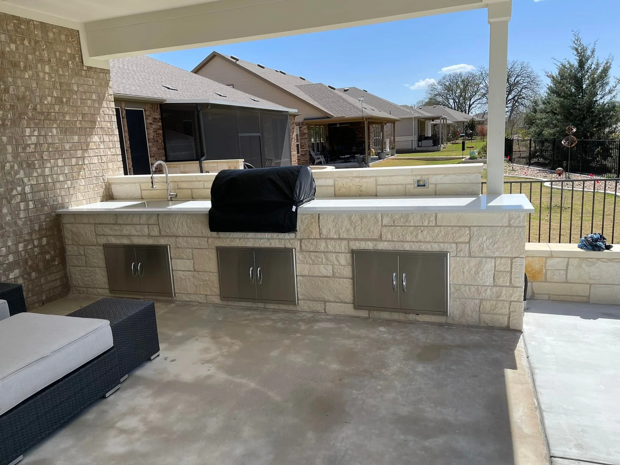 Outdoor kitchen with a built-in grill, sink, and refrigerator under a covered patio, overlooking a backyard with a lawn, trees, and a black fence.