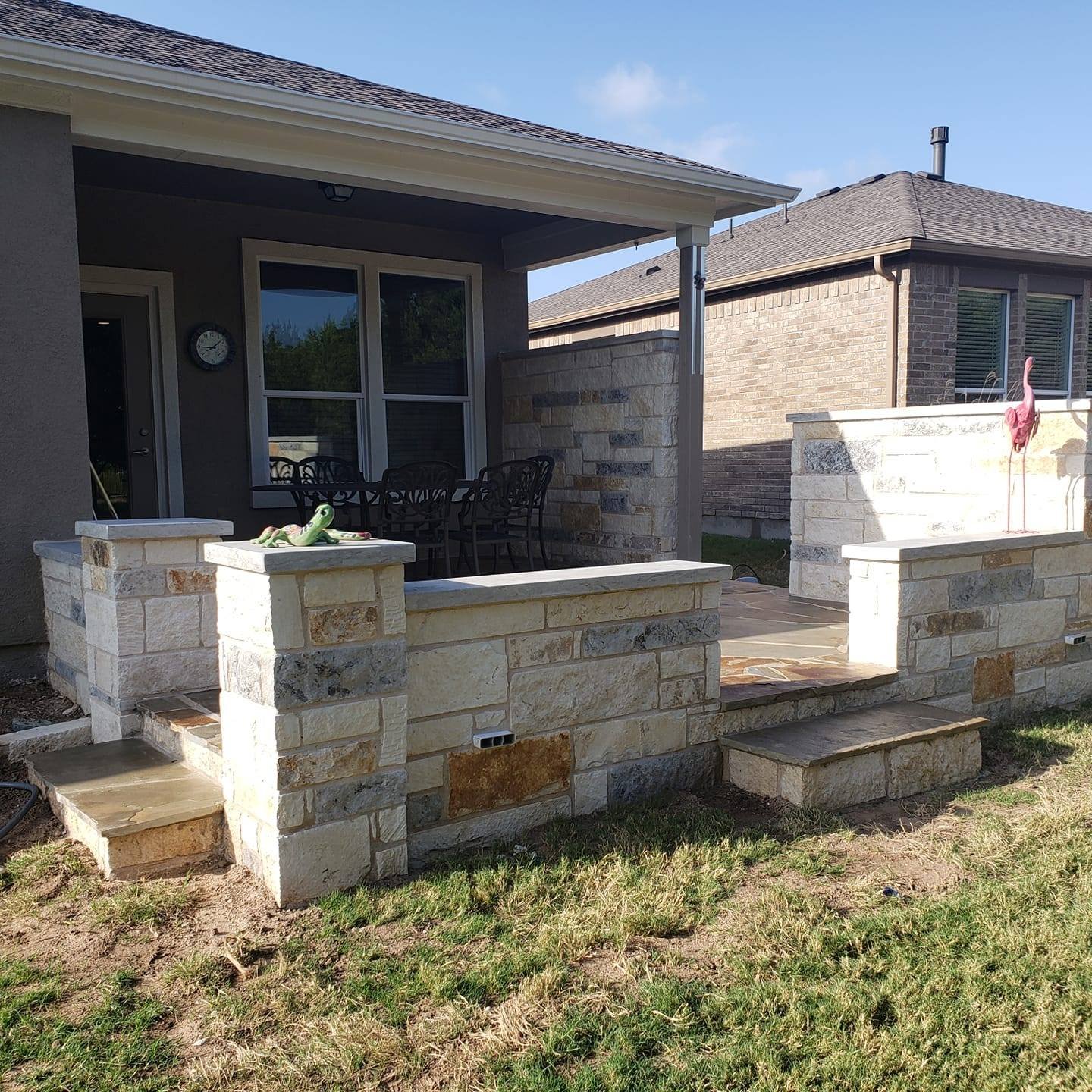 Backyard patio with a stone retaining wall, stairs, and a covered sitting area with a dining table and chairs. A decorative flamingo and a garden decoration on top of the wall. House walls are brick and stucco, and the sky is clear.