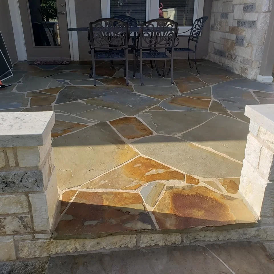 A stone patio with a raised step leading to an outdoor dining area with a black metal table and chairs, and a glass door in the background.