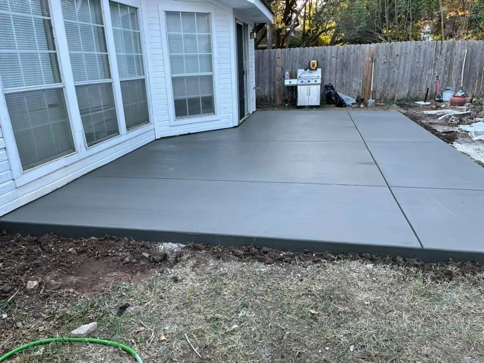 Freshly poured concrete patio adjacent to a house with large windows, enclosed by a wooden fence, with patio tools and barbecue grill in the background.