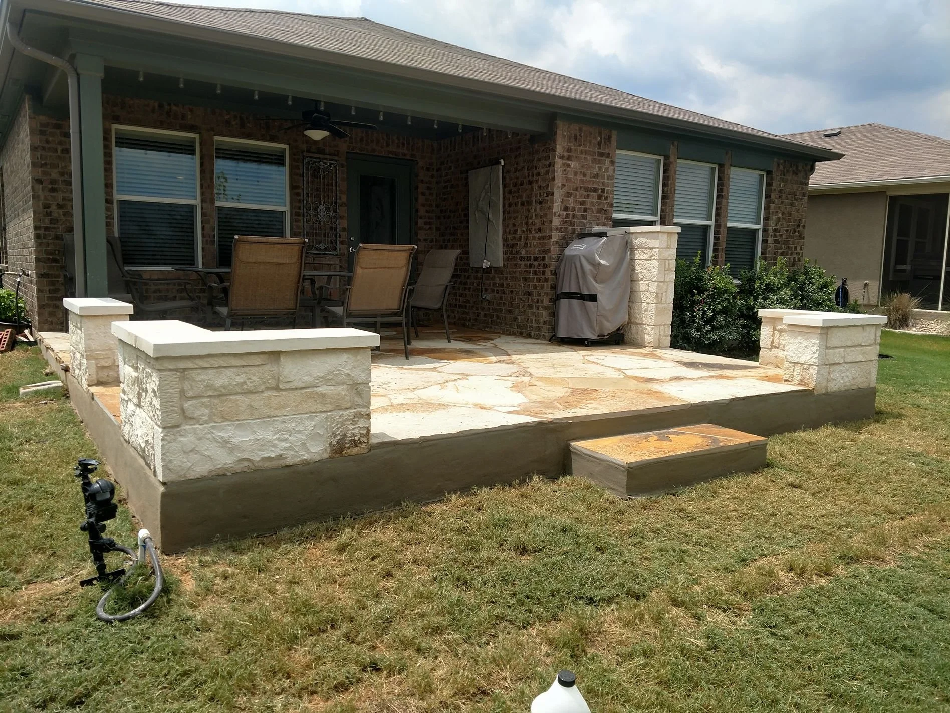 Backyard patio with a stone deck, outdoor dining table with chairs, patio cover, a grill, and brick house in the background.