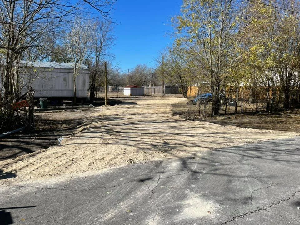 A dirt driveway leading to a backyard with trees, a shed, and a fence, under a clear blue sky.