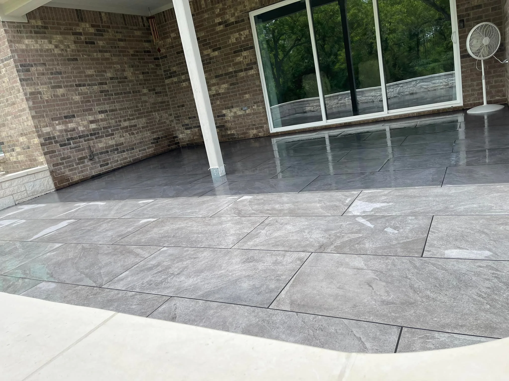 This image shows an outdoor patio area with large gray tiles, a glass sliding door, a brick wall, and a white fan. The patio appears to be wet.