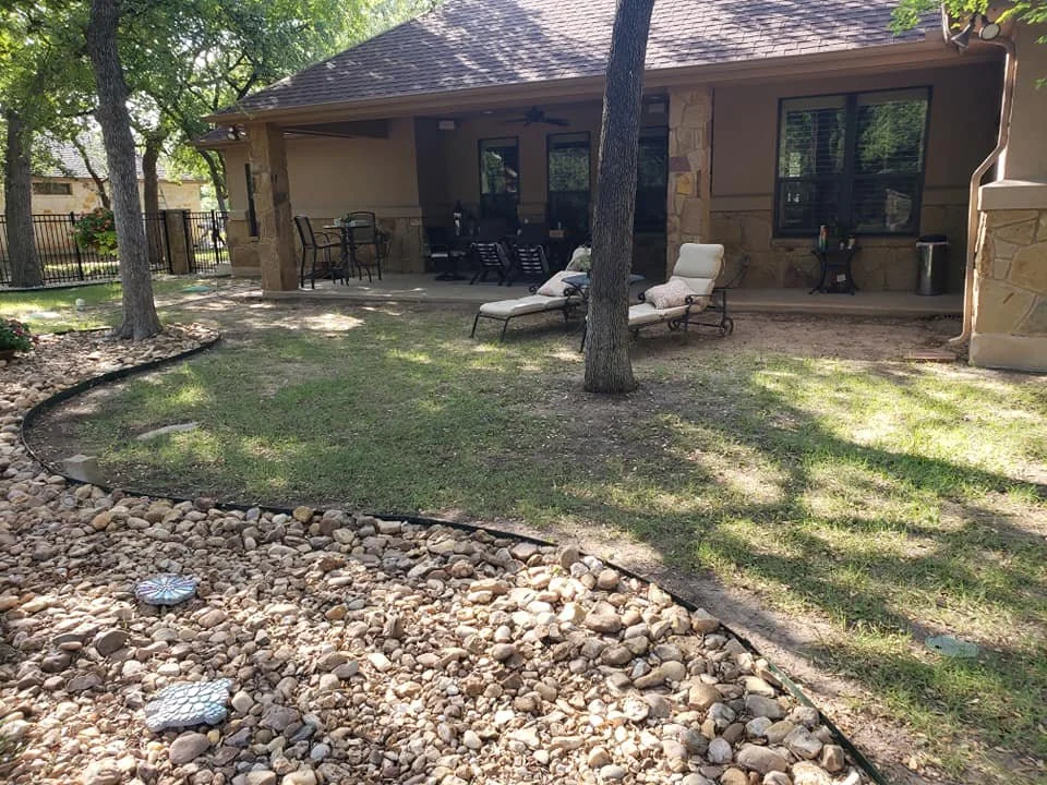 Backyard patio with outdoor seating, lounge chairs, and a table, surrounded by trees and rock landscaping.