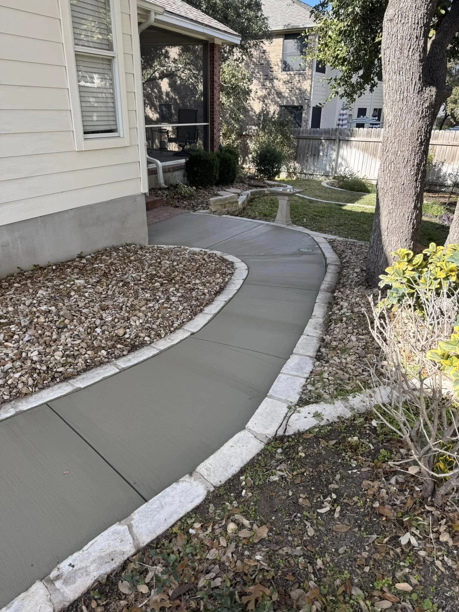 A curved concrete walkway bordered by stone edging, running alongside a house with a screened porch in a backyard. The yard has trees, bushes, and a garden area, with a bench and some landscaping features.
