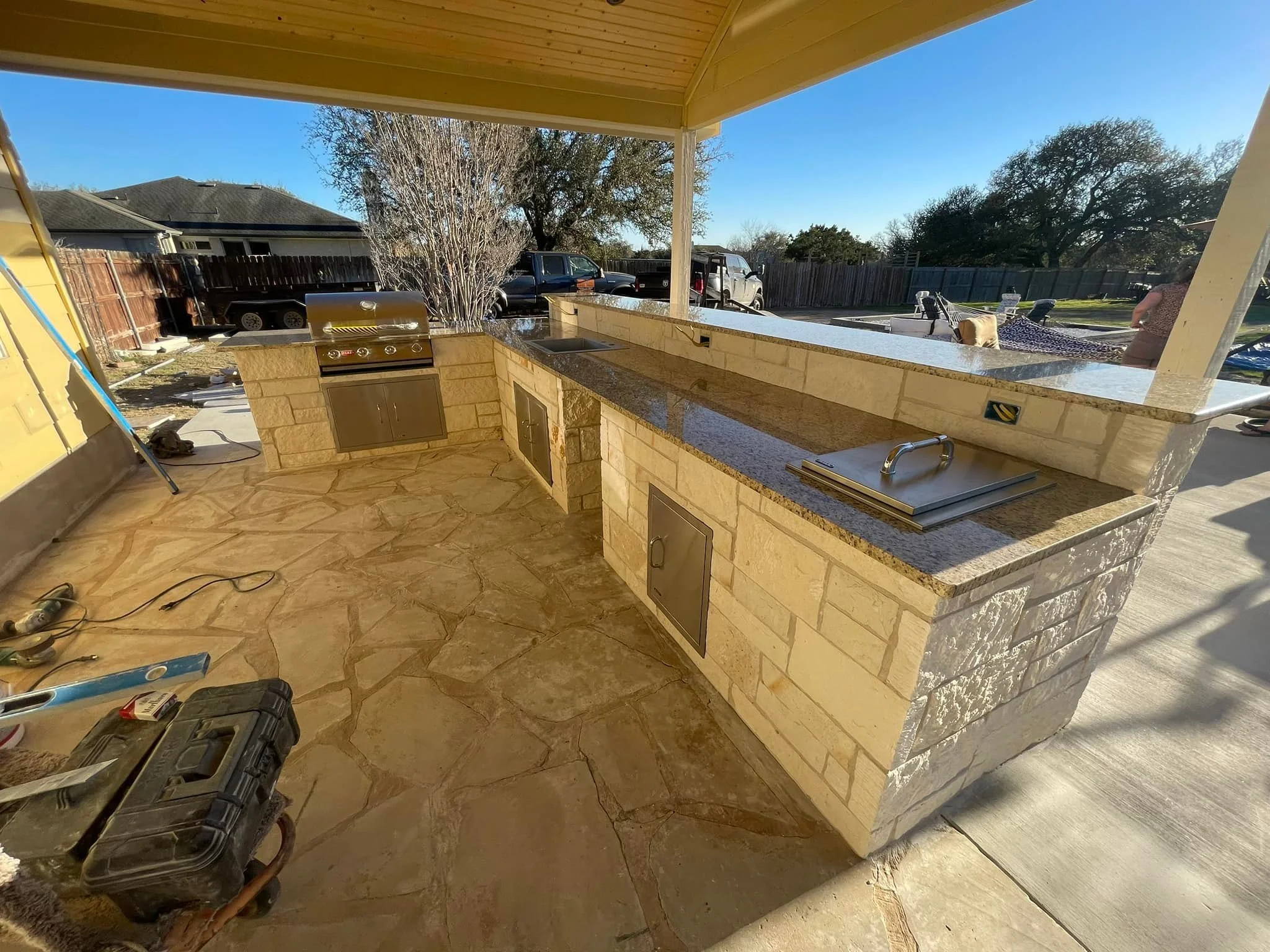 An outdoor kitchen under construction with a stone counter, built-in grill, sink, and storage cabinets. There are construction tools and equipment on the floor, and a wooden roof overhead. In the background, there are parked cars, trees, and a fenced