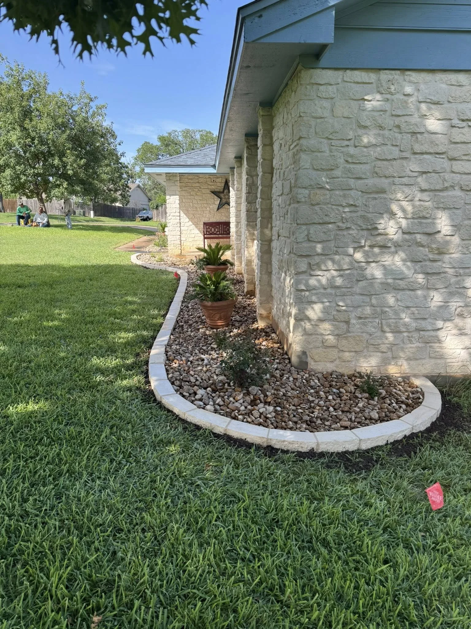 A landscaped yard next to a house with a stone exterior, featuring a curved bed with decorative stones, potted plants, and a garden bench. In the background, a lawn, trees, and a few people are visible.