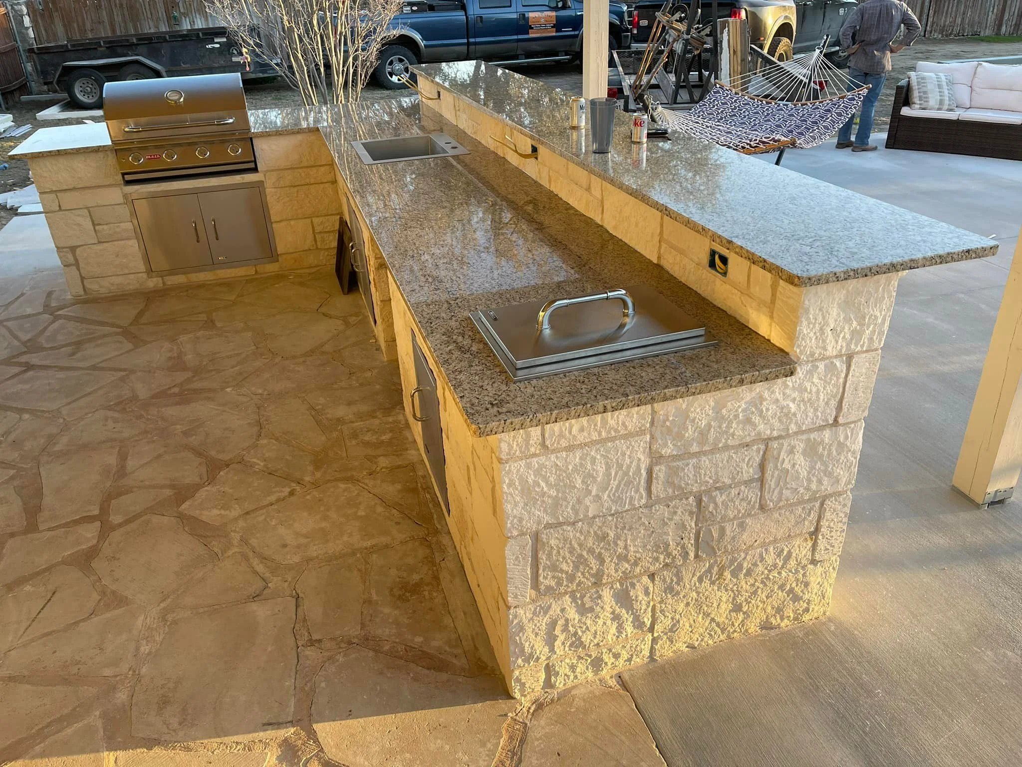 Outdoor kitchen with stone countertops, built-in grill, and a small sink, with a stone and granite structure, and outdoor furniture including a hammock and seating area in the background.