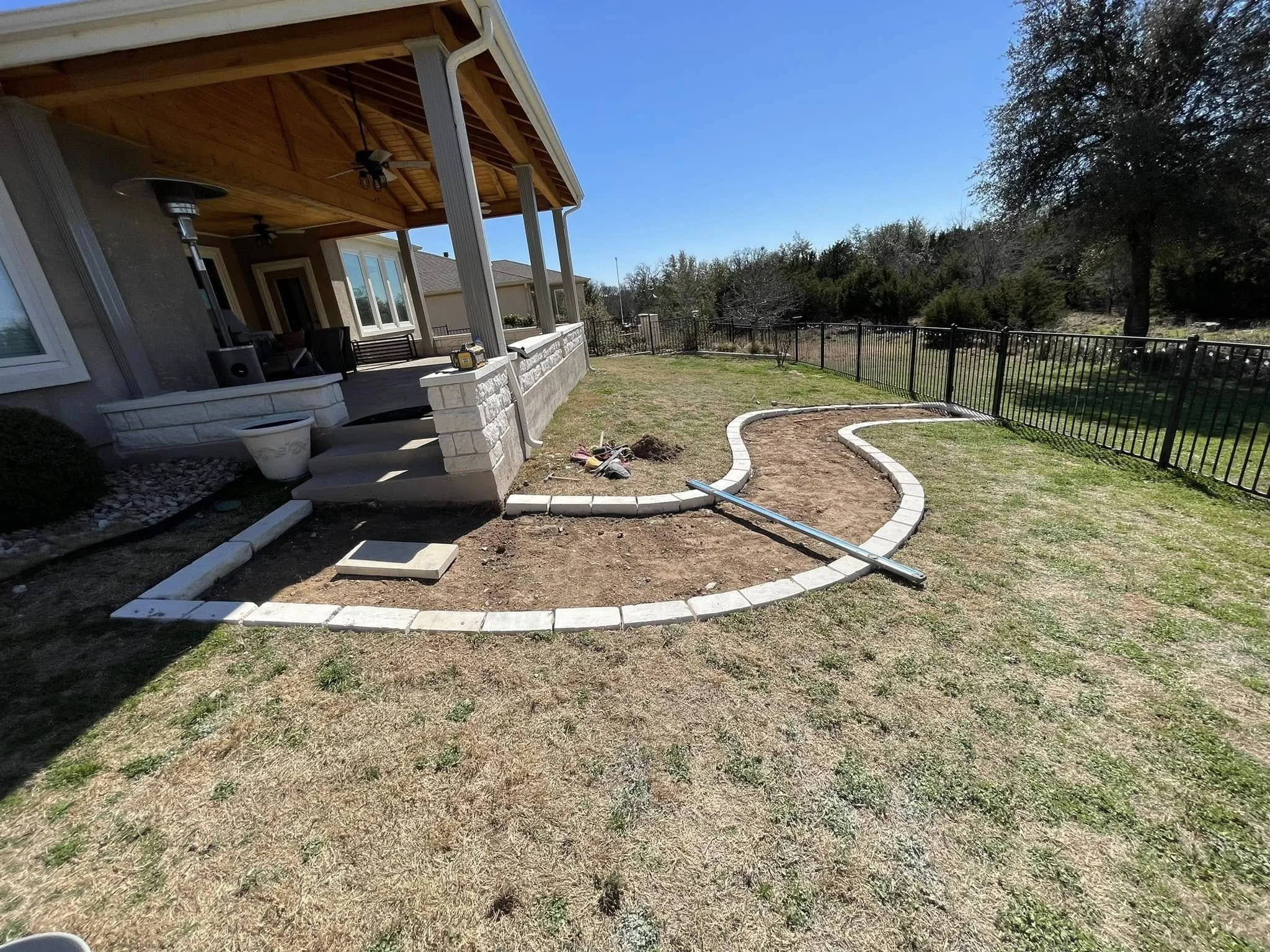 Backyard patio area under construction with curved stone border for landscaping, some soil prepared, and tools present.