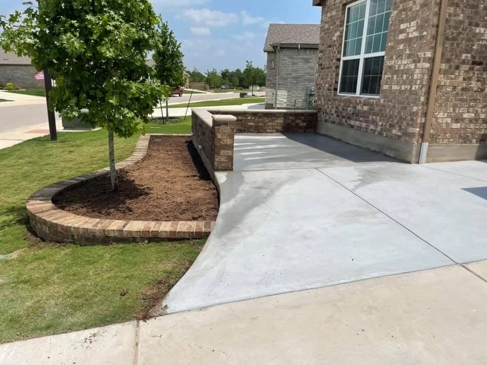 A brick garden bed with a small green tree next to a house. The garden bed has freshly turned soil. There is a concrete sidewalk and driveway in front of the house, with part of a brick wall and large window visible.