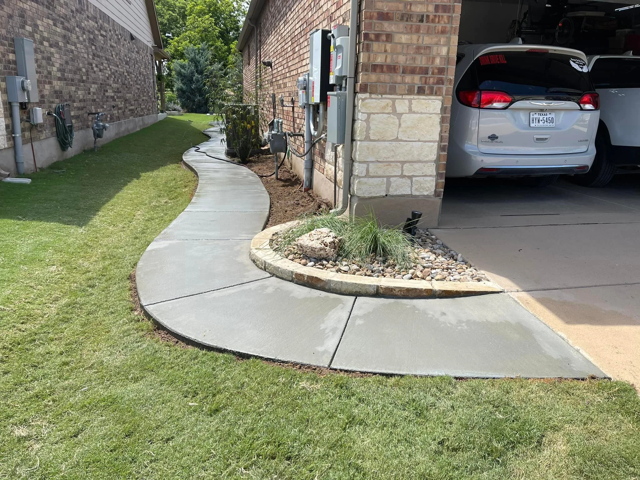 Curved concrete sidewalk alongside a house and a garage, with a landscaped area containing rocks and plants at the corner.
