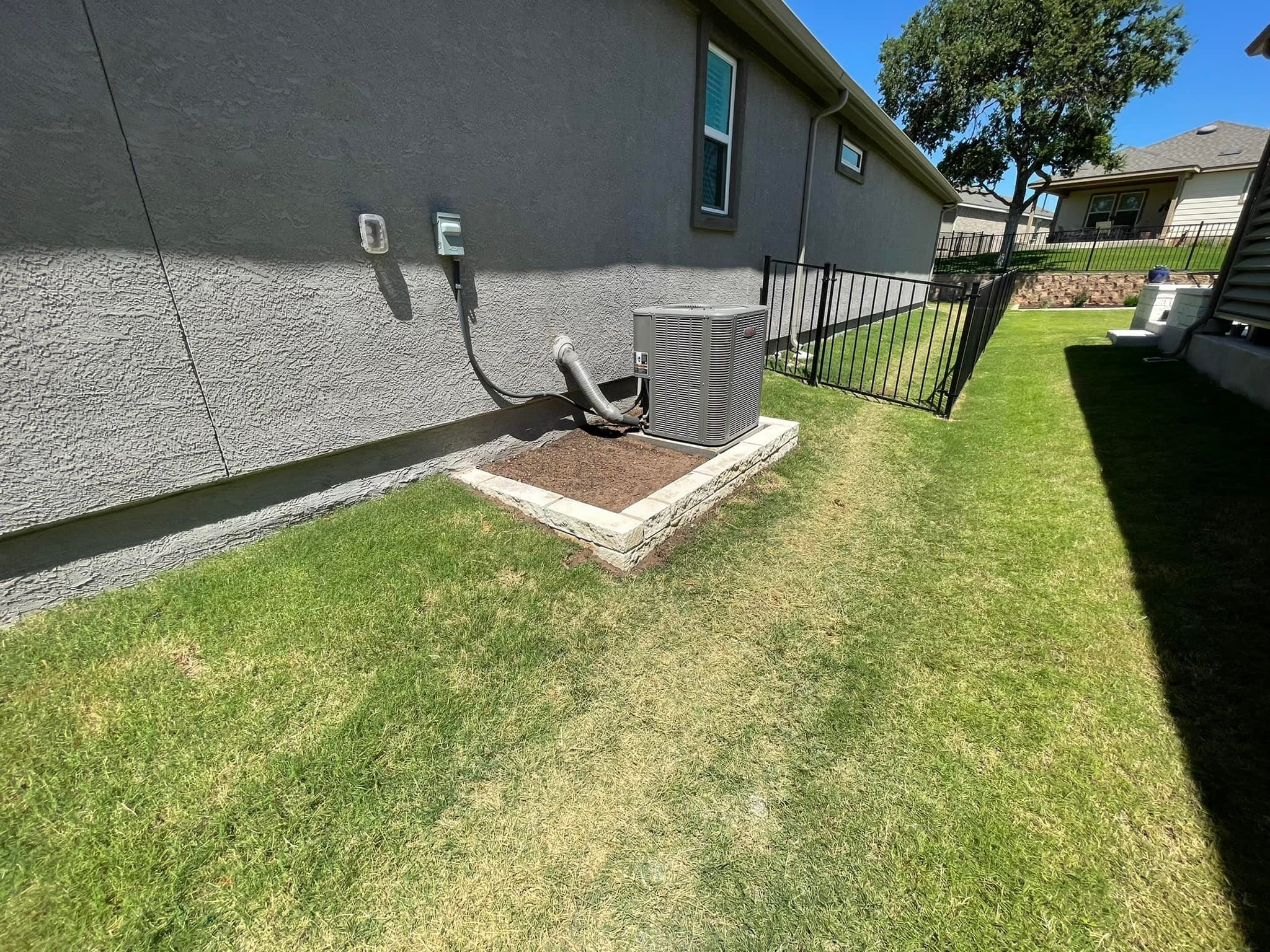 Residential backyard with lawn, gray house wall, HVAC unit on concrete slab, small garden bed, and black metal fence, with neighboring houses in the background.