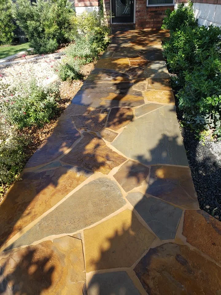 Stone pathway leading to a house entrance, bordered by bushes and plants on both sides, with shadows cast from nearby trees.