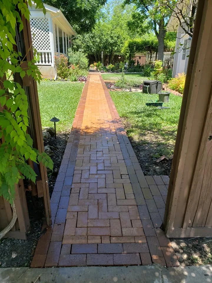 A brick pathway running through a lush green backyard with trees, shrubs, and a white porch on the left. The pathway is bordered by grass and flower beds, extending towards a fenced area in the distance under a clear blue sky.