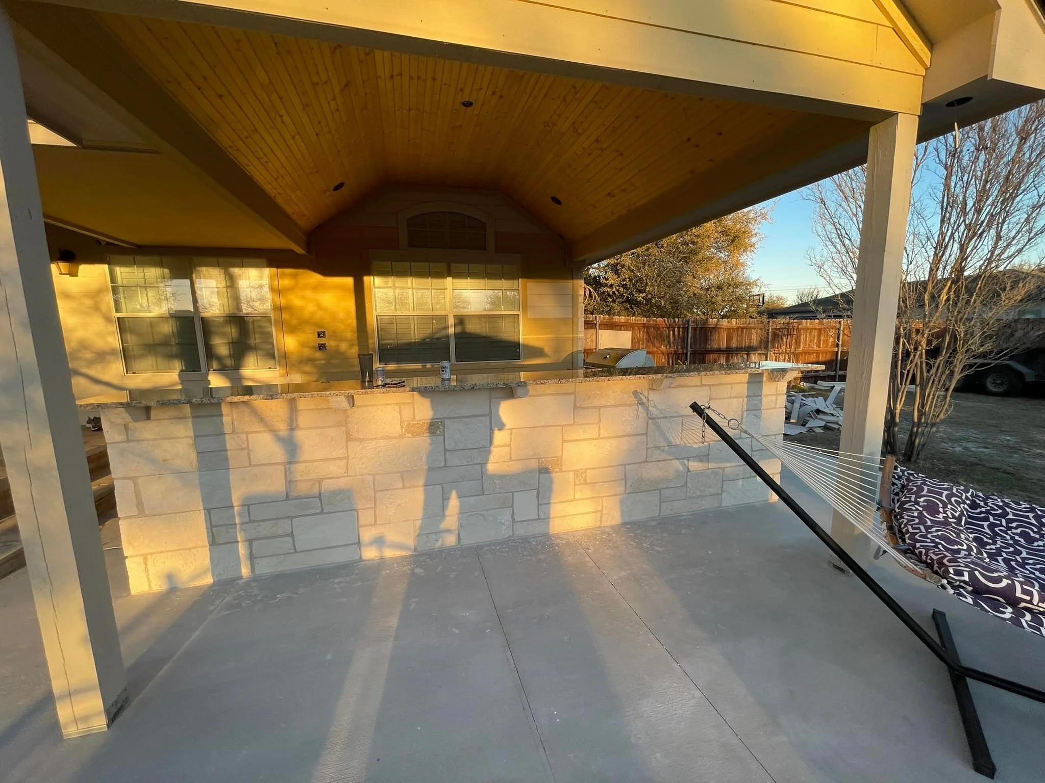 Back porch of a house at sunset, with a stone counter, wooden roof, and a hammock on the right side, with trees and a fence in the background.