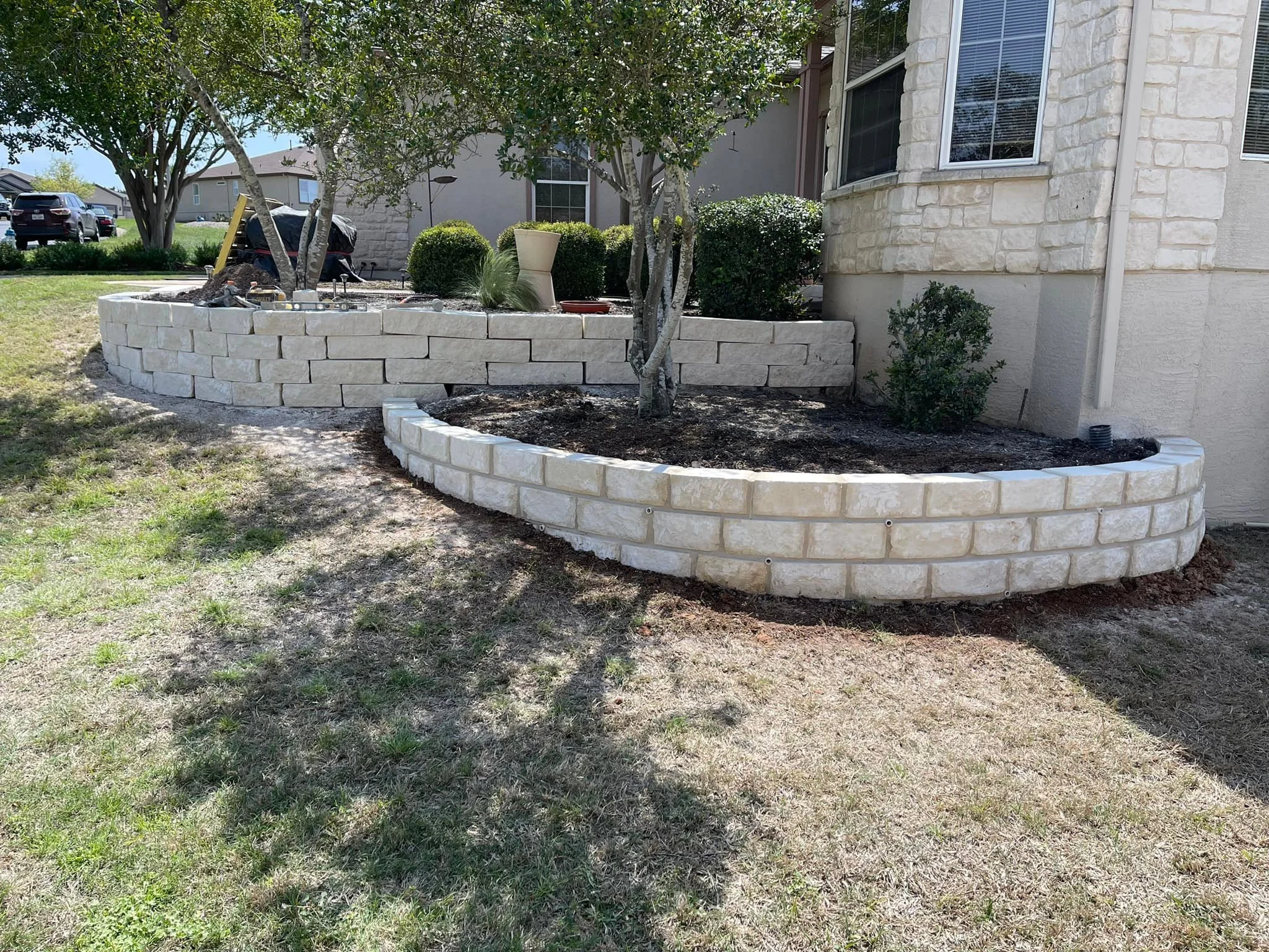 A landscaped backyard with a curved stone retaining wall and a tree in the center. The wall has light-colored stones with soil and small shrubs behind it. The house exterior with windows and a garden bed with potted plants are visible in the backgrou