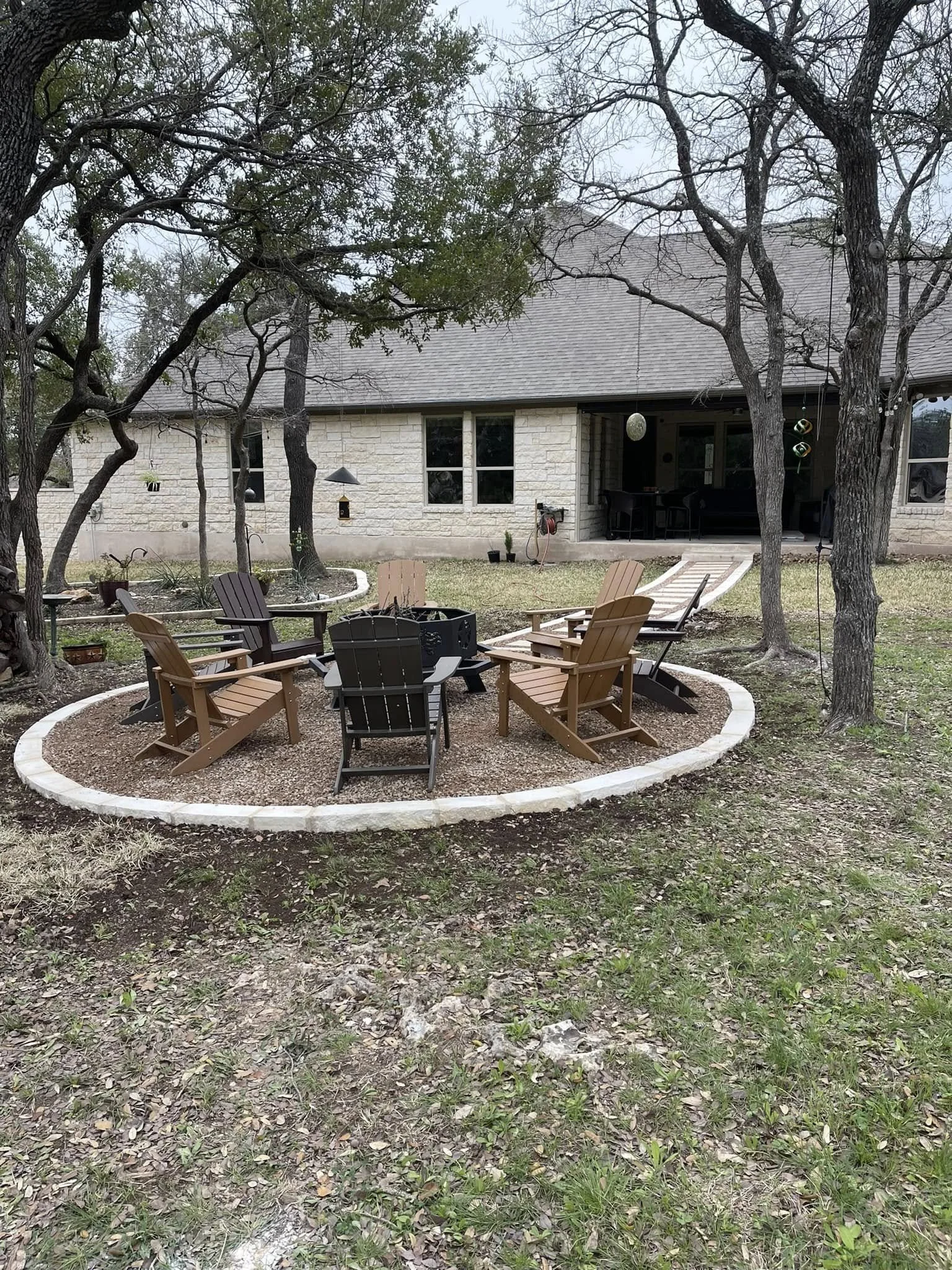 A backyard with a circular fire pit area surrounded by Adirondack chairs and two other chairs, with a house in the background and trees around the yard.