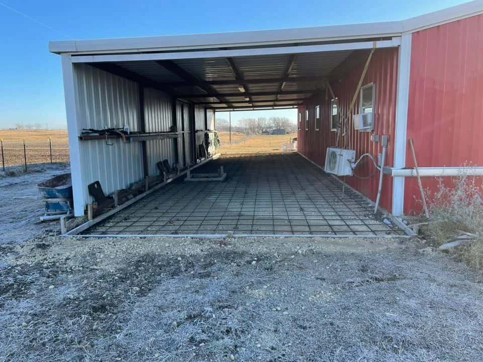 Open-sided metal shelter with a concrete slab foundation, attached to a red building, with a gravel ground in the foreground.