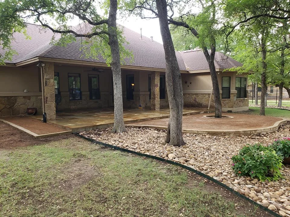 Backyard with large stone house, covered patio, trees, landscaped area with rocks, mulch, and a flower bush.