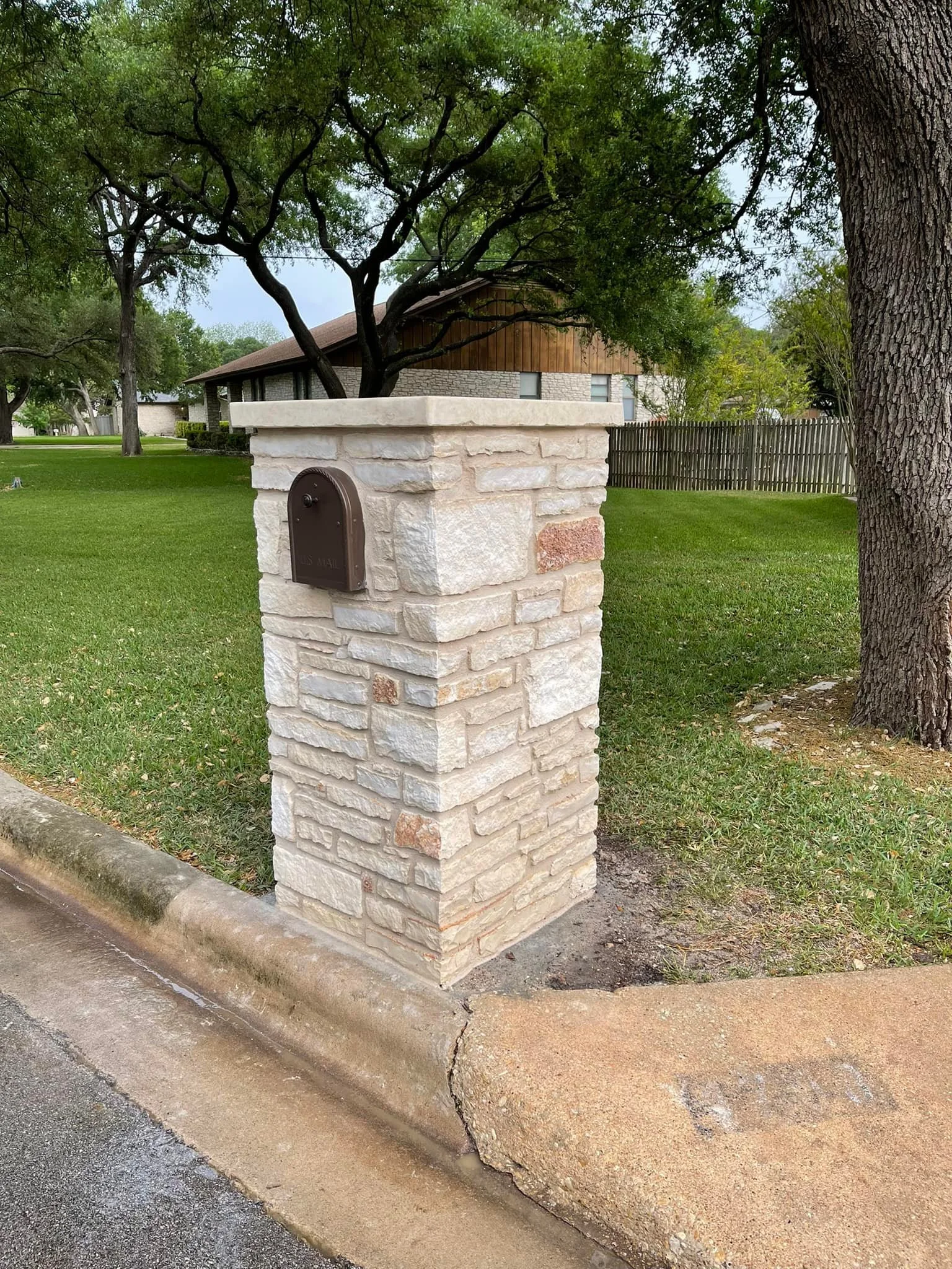 A stone mailbox on a sidewalk next to a grassy yard with trees and a house in the background.