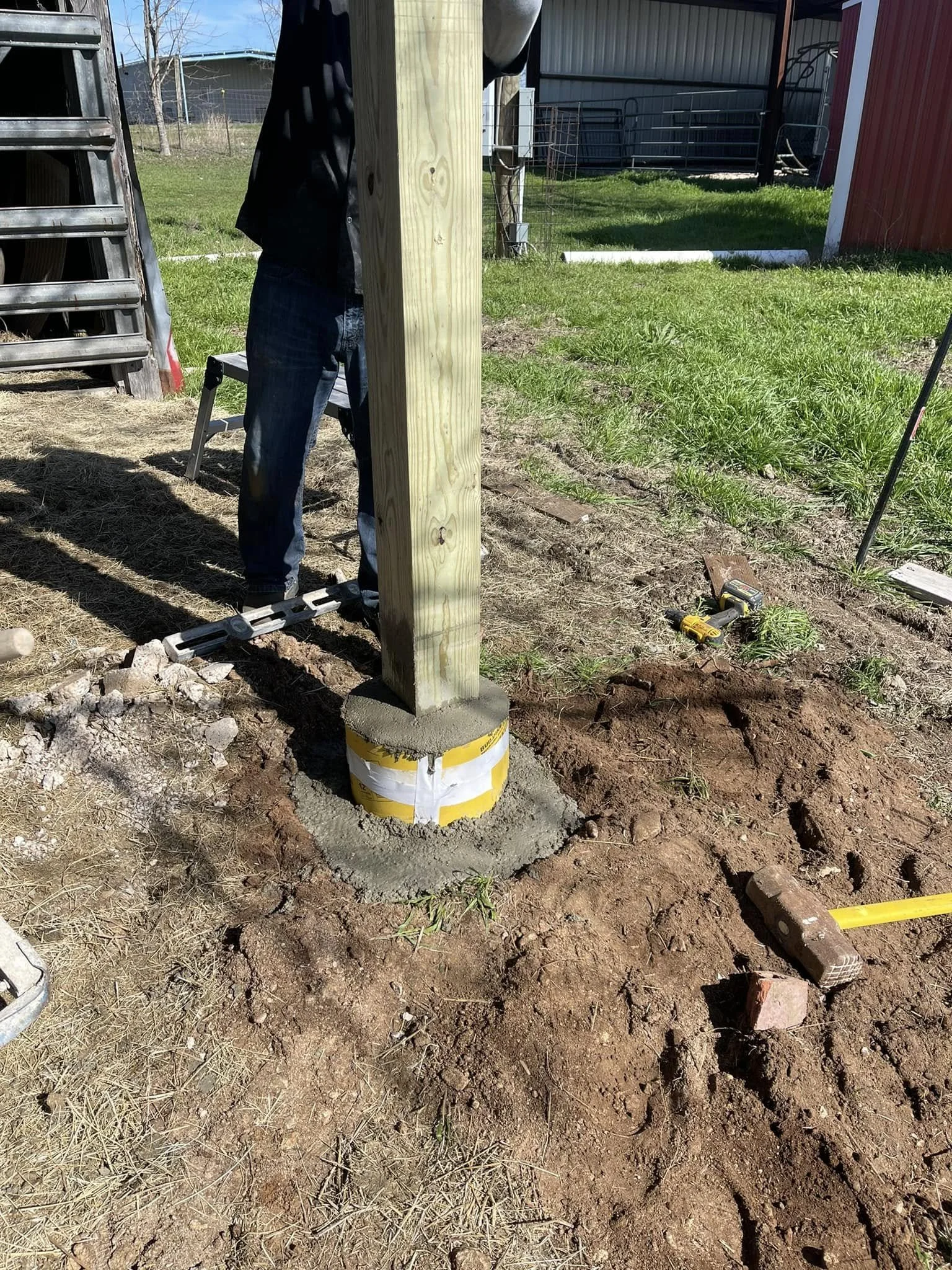 A construction site outdoors with a wooden post set on a concrete footing, surrounded by dirt and tools, including a yellow measuring tape and a cordless drill. Part of a person wearing jeans and a black jacket is visible in the background.