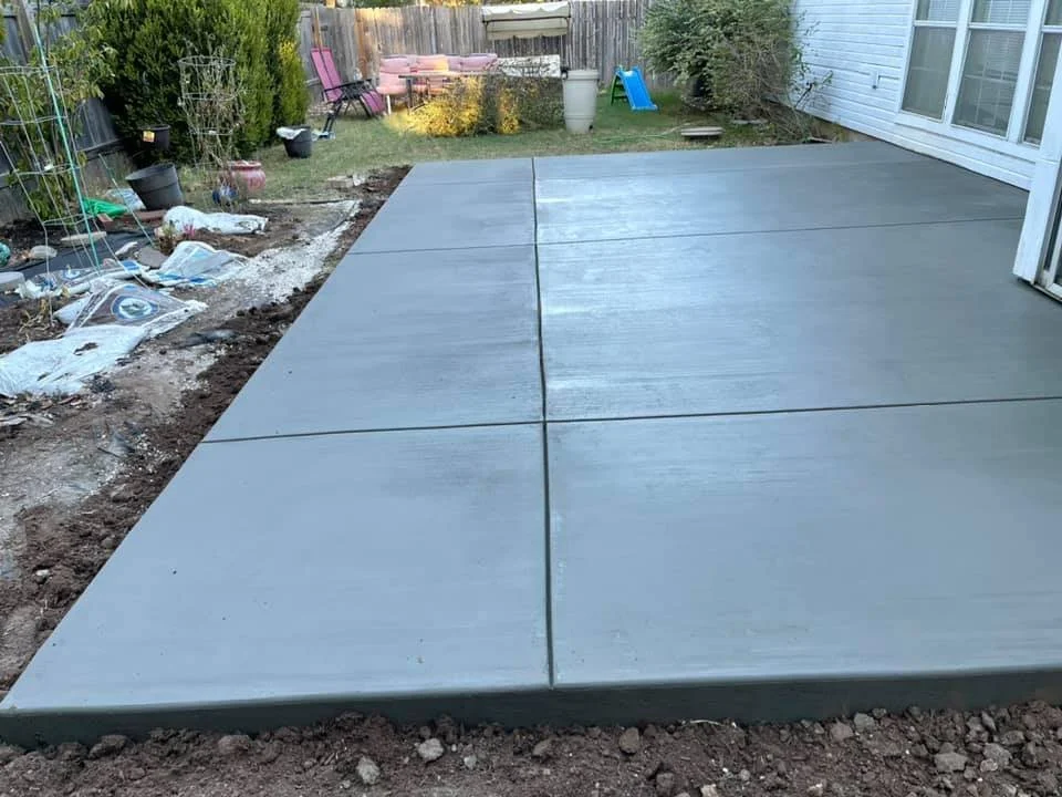 Newly poured concrete patio with large square slabs in a backyard, surrounded by a wooden fence, garden plants, outdoor furniture, and a sliding glass door on the house.