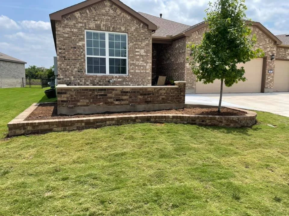 Front yard of a brick house with a small tree in a landscaped bed, a white window, and a concrete driveway.