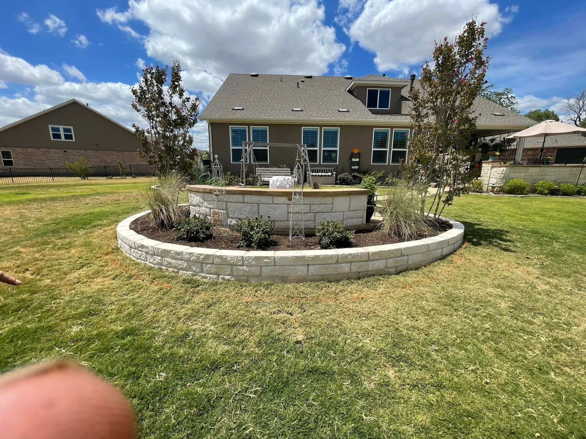 View of a backyard with a stone raised flower bed with plants and trees, in front of a gray house with large windows, under a partly cloudy sky.