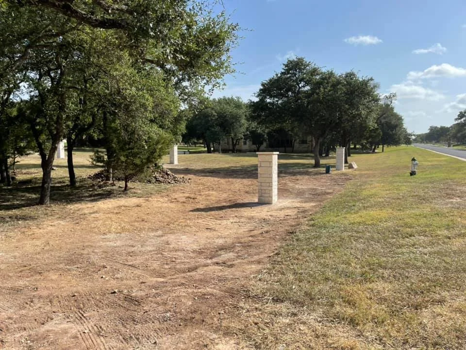 A rural scene with dirt ground, grass, trees, brick pillars, and a paved road on the right under a partly cloudy sky.