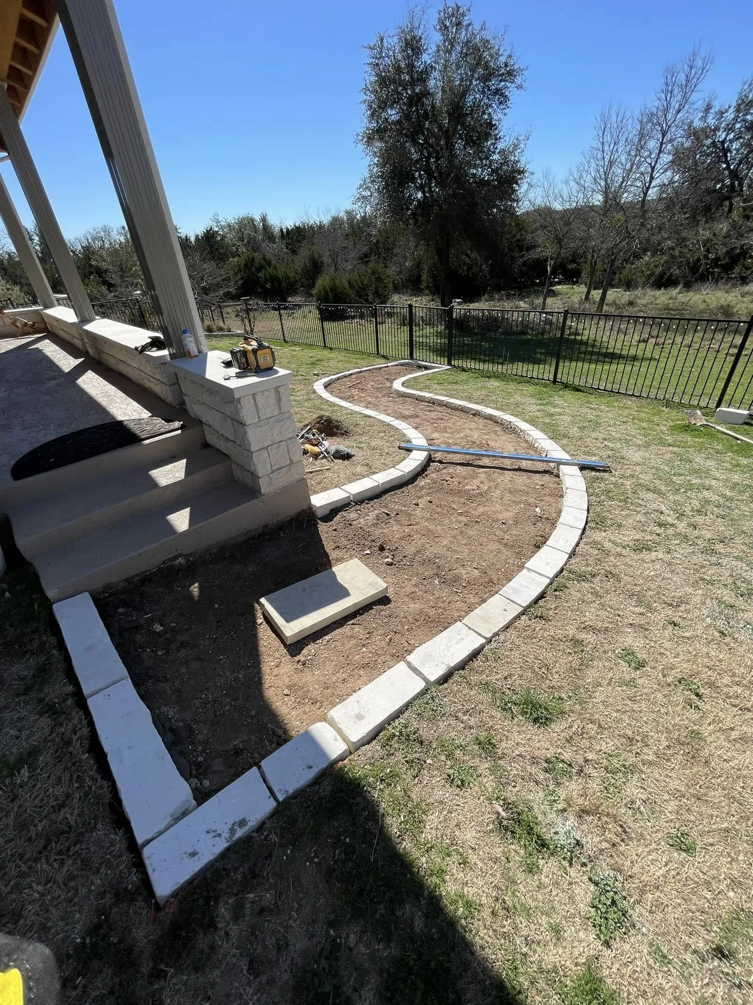 A backyard with a curved walkway under construction, bordered by white bricks, with a partially built stone porch and some construction tools on the stairs.