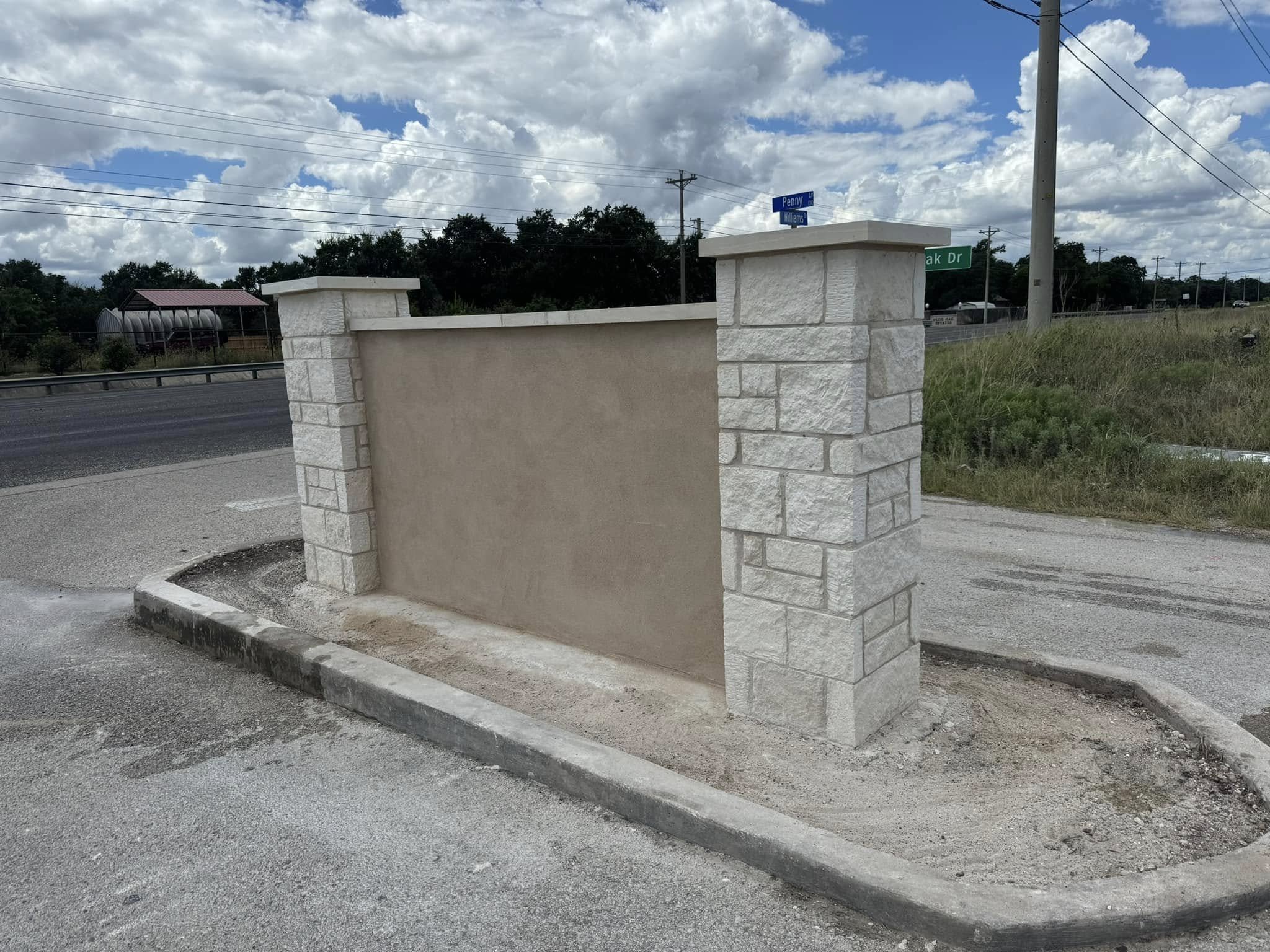 A beige wall with stone pillars on each side, located at the edge of a parking lot near a street, with street signs visible in the background against a partly cloudy sky.