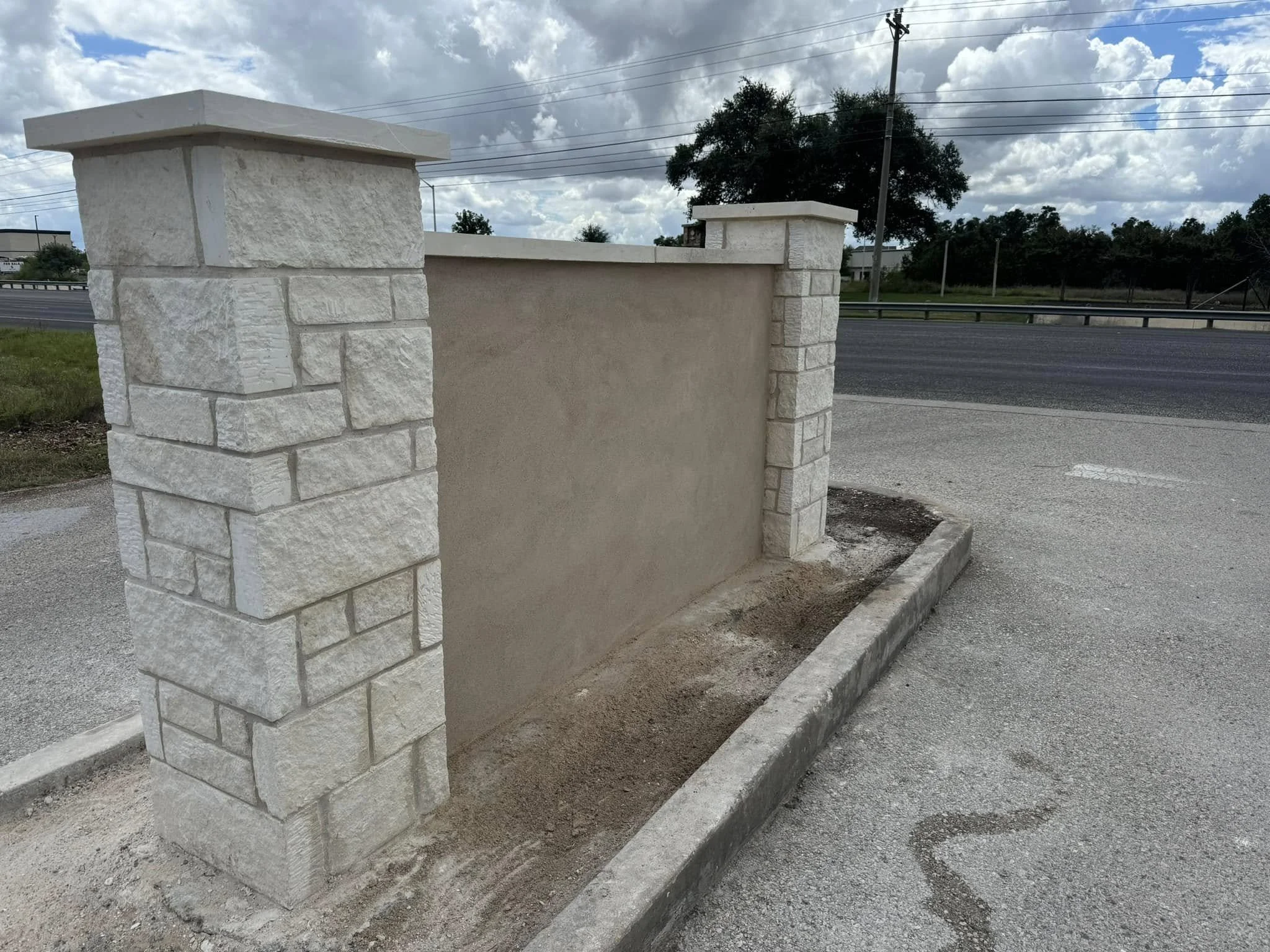 A partially built decorative wall with stone columns and a stucco wall underneath a cloudy sky, next to a paved road and utility poles.