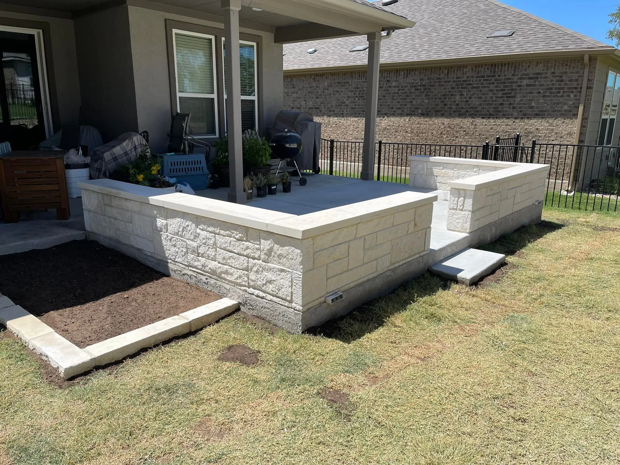 Backyard patio with a stone wall and a small set of stairs leading up to it. The patio is partially covered by a roof and has outdoor furniture, a grill, and potted plants. There is a grassy yard area with a small garden bed in the foreground.