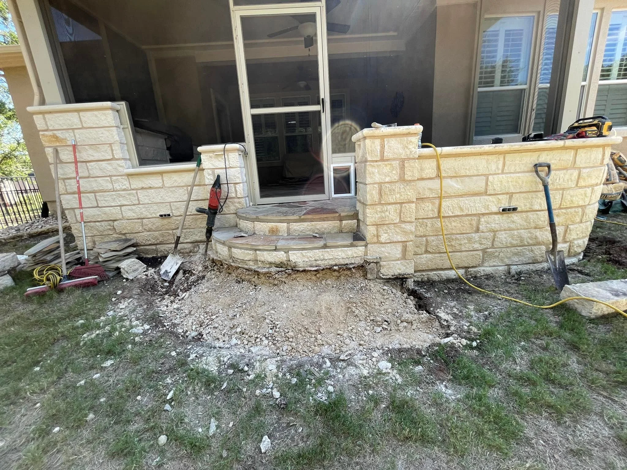 Construction site with stone steps and a low stone wall around a porch, with tools and materials scattered around, and a partially excavated area in front of the steps.