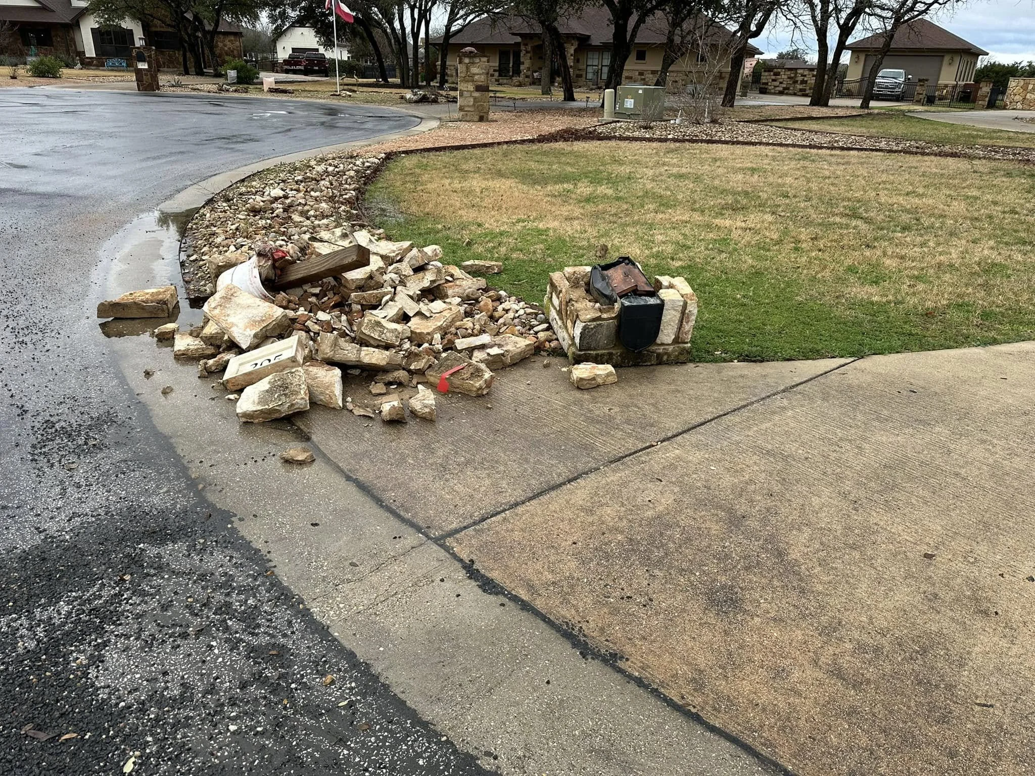 Debris from a collapsed brick wall and sidewalk on a residential street corner, with a pile of rocks, bricks, and construction supplies next to a brick post, and an intact sidewalk and grassy yard in the background.