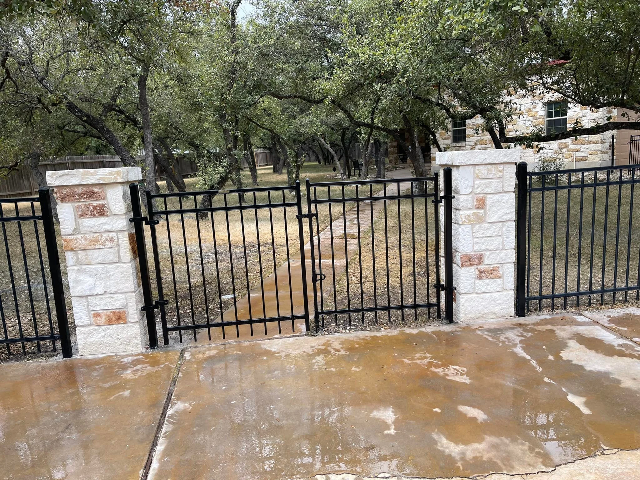 Black metal gate with vertical bars and latch, flanked by white brick pillars, leading to a backyard with trees and a stone house in the background. Wet concrete porch in foreground.