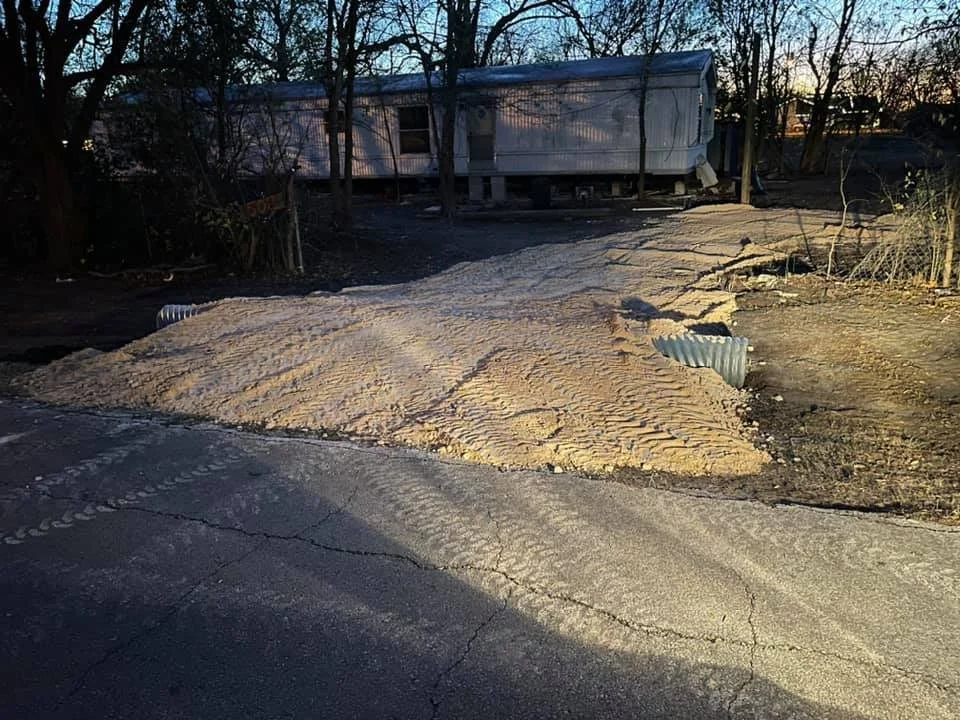 A trailer on a raised foundation with sand piled in front, partially covered by a tarp, next to a cracked driveway during sunset.
